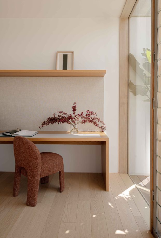 A warm, minimalist study nook with a built-in timber desk and raised ledge, a textured upholstered wall panel, and a rust-brown upholstered chair. Pale timber flooring and matching timber window framing sit beside full-height glazing, with soft sunlight casting shadows across the floor. A small framed artwork and vase of branches provide restrained decoration.