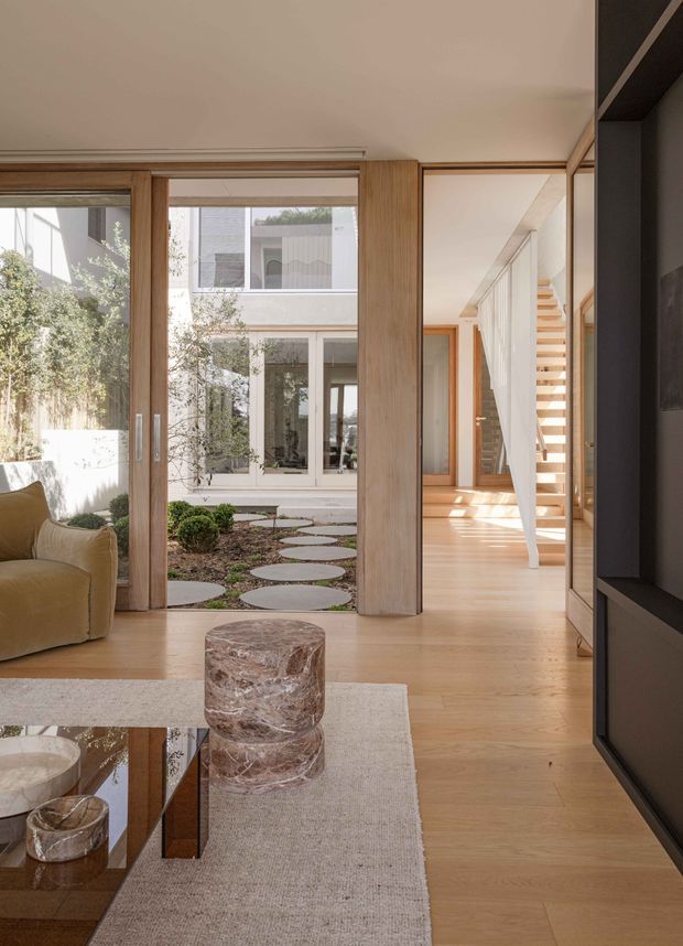 A bright, refined contemporary living room opening onto a small landscaped courtyard. The space features pale timber flooring, extensive timber-framed glazing and sliding doors, a dark built-in media wall, a low reflective coffee table, a textured neutral rug, and a sculptural stone-like cylindrical stool or side table. A timber staircase and light-filled hallway are visible beyond the courtyard connection.