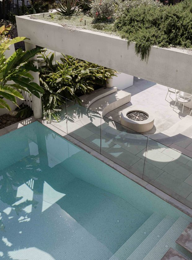 A sunlit contemporary outdoor courtyard viewed from above, featuring a turquoise tiled swimming pool, glass pool fencing, pale stone or concrete paving, a raised concrete planter with dense tropical and succulent planting, built-in bench seating, and a circular built-in fire or planter feature. The space has clean geometric forms, abundant greenery, and strong afternoon shadows.