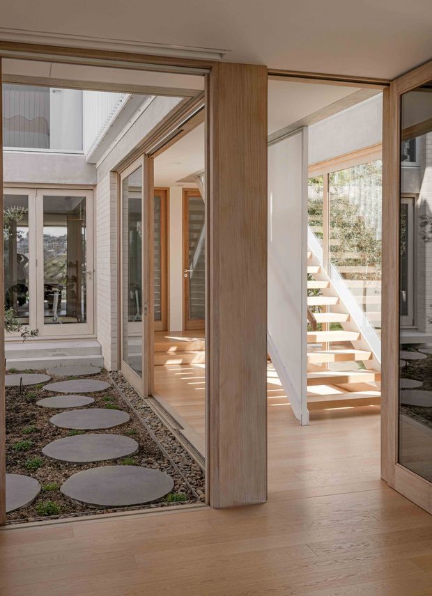 A bright contemporary interior corridor opening onto a landscaped courtyard, with extensive timber-framed glazing, pale timber flooring, white walls, and an open-riser staircase. Circular concrete stepping stones and gravel landscaping are visible through the glass, while natural light filters through the courtyard and upper-level openings.