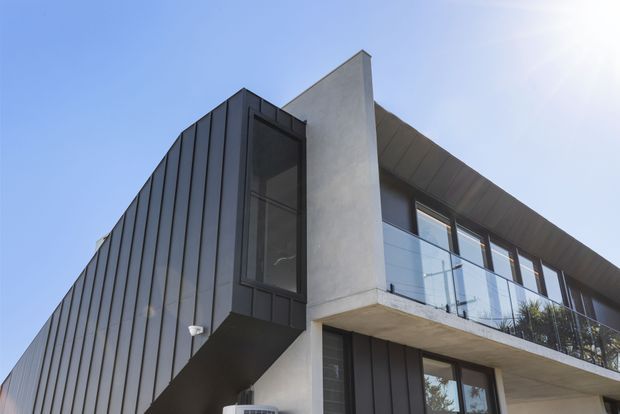 Low-angle exterior view of a contemporary multi-storey residence with dark vertically ribbed cladding, a sharply projecting upper window volume, a pale rendered or concrete central wall, and a cantilevered balcony with clear glass balustrade. The building has a strong angular composition and a broad sloping or pitched roof form against a clear blue sky.