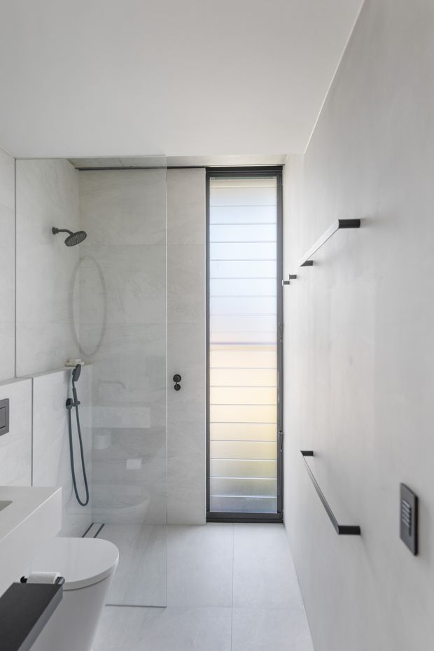 A compact contemporary bathroom with a walk-in glass shower, wall-mounted dark shower fittings, a white toilet and integrated basin, pale large-format floor and wall tiles, and a tall translucent louvred-style window. Minimal black grab rails and fixtures contrast with the light neutral finishes.