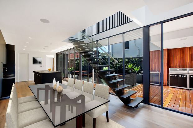 Bright contemporary open-plan dining area with a large glass-topped dining table and upholstered chairs. A dramatic floating timber staircase with black metal framing rises through a glazed atrium, with indoor planting beneath. Through the glass partition is a timber-lined outdoor entertaining area with built-in barbecue cabinetry. The space has white walls, light timber flooring, dark cabinetry and extensive glazing.