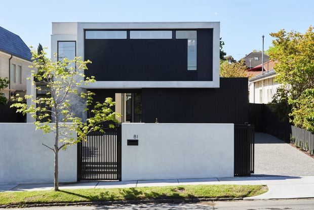 Street-facing view of a contemporary two-storey residence with a minimalist rectilinear form, flat roof, white rendered walls, dark vertical cladding, and large dark-framed glazing. A rendered front boundary wall, black metal slat pedestrian gate, driveway, and landscaped verge define the frontage.