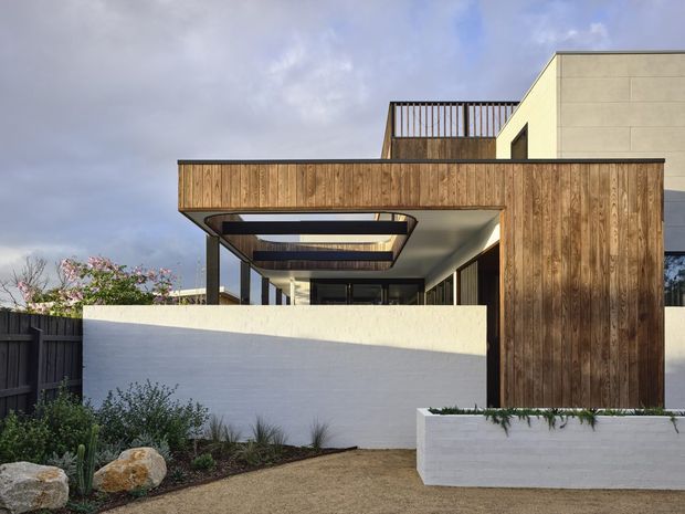 Contemporary multi-level residential facade with warm vertical timber cladding, pale rendered masonry walls, projecting upper volumes, and a covered outdoor terrace or entry zone. The composition features a broad timber-clad horizontal canopy, dark slender structural posts, recessed ceiling lighting, and rooftop or upper-level balustrading. Low landscaped planting and rendered boundary walls frame the foreground.