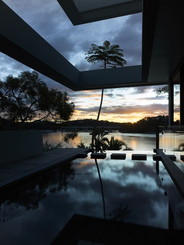 A dramatic dusk view from a covered outdoor terrace overlooking a calm lake or river. The foreground features a dark reflective pool or water feature, timber decking, concrete or rendered walls, glass balustrades, and a large cantilevered roof structure with an open skylight. Silhouetted palms and native trees frame the sunset and are reflected in the water, creating a moody, tranquil atmosphere.