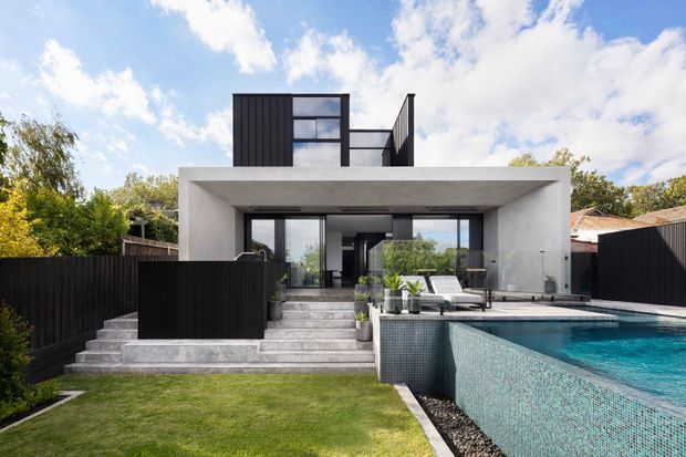 Contemporary two-storey residence with a stark rectangular concrete frame, dark vertical cladding, extensive glazing and a stepped terrace leading to a turquoise tiled swimming pool. The landscaped backyard includes lawn, planting and black privacy fencing under a bright partly cloudy sky.