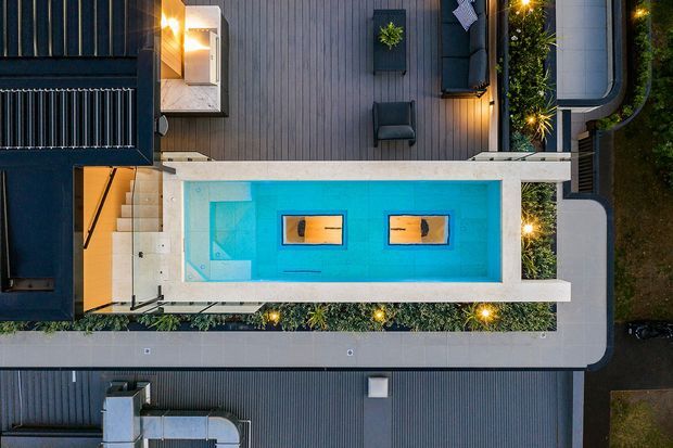 Overhead aerial view of a contemporary residential outdoor area, centred on a long rectangular swimming pool with bright turquoise water and two in-pool sun loungers. The pool is surrounded by pale concrete paving, dark timber or metal-clad building surfaces, planted garden strips, outdoor seating, stair access, and warm integrated landscape lighting. The composition strongly emphasizes the pool terrace and modern geometric site layout.