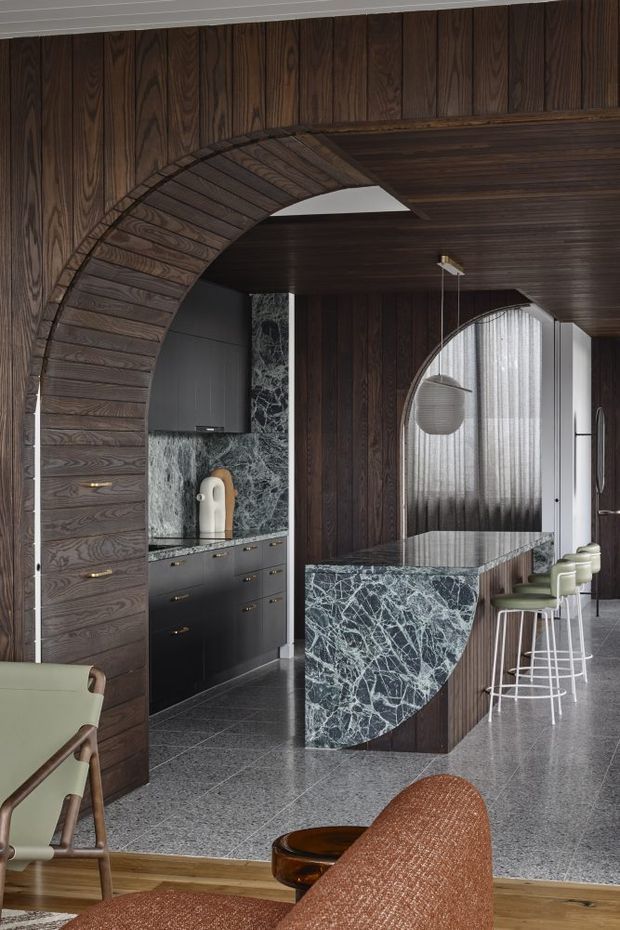 A sophisticated open-plan kitchen and living area featuring dark timber wall and ceiling panelling, a dramatic curved timber archway, dark cabinetry, and a veined dark stone splashback and island. The island has a sculptural curved end and is paired with pale upholstered bar stools. Terrazzo-style flooring transitions to timber flooring in the foreground, with a pendant light and sheer-curtained arched glazing adding softness.