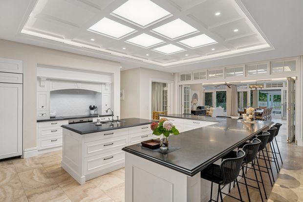 A spacious, bright traditional kitchen with two large white islands, dark stone benchtops, extensive white cabinetry, integrated appliances, and a tiled floor. The kitchen opens through glazed doors into an adjoining dining or living area. A highly detailed coffered ceiling with multiple illuminated skylight-style panels and recessed lighting forms a prominent architectural feature.