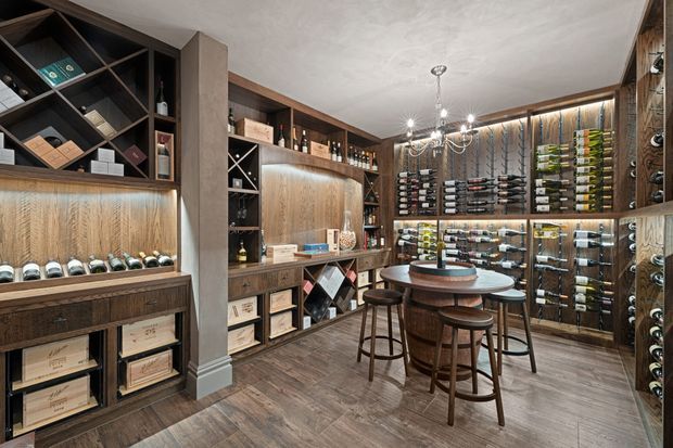 A sophisticated residential wine room with extensive dark timber wall-mounted wine storage, illuminated bottle displays, built-in cabinetry, a central round tasting table, and tall stools. The space has warm layered lighting, wood-look flooring, and a refined cellar-inspired atmosphere.