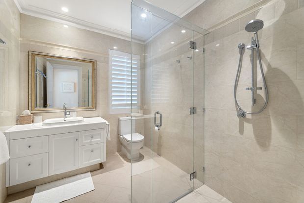 Bright, refined bathroom featuring a white double-sink vanity with framed mirror, wall-mounted toilet, and a large frameless glass walk-in shower with dual shower fixtures. Beige stone-look wall and floor tiles, white plantation shutters, recessed ceiling lights, and subtle traditional detailing create a clean, upscale interior.