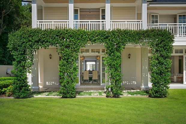Front exterior view of a refined two-storey residence with a symmetrical covered veranda, white square columns, white balustrades, pale horizontal weatherboard cladding, and climbing greenery framing the ground-floor openings. French doors and a glimpse of a dining area are visible beneath the veranda, with a manicured lawn in the foreground.