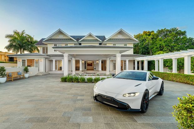 A large, light-coloured traditional-style residence viewed from the front, featuring multiple gabled roof sections, a broad classical portico with columns, and an expansive paved driveway or forecourt. A white sports car occupies the foreground. A covered pergola-like walkway extends along the right side, with tropical landscaping and hedges around the property.