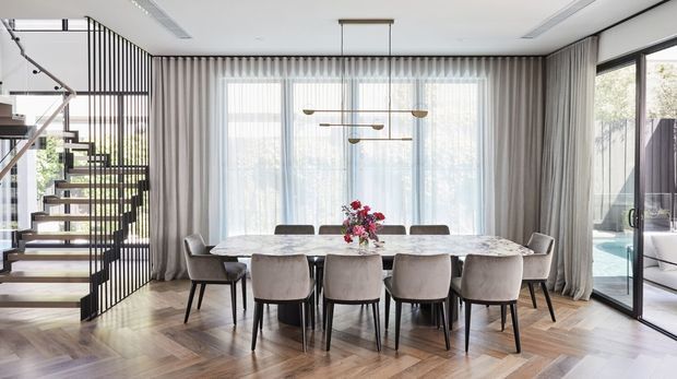 A bright, refined dining area with a large marble-topped dining table surrounded by upholstered chairs. Full-height sheer and draped curtains frame expansive glazing, while a sculptural linear pendant hangs overhead. A floating timber staircase with black metal balustrading is visible to the left, and a glazed door opens toward an outdoor pool area on the right. Warm herringbone timber flooring and neutral furnishings create an elegant contemporary interior.