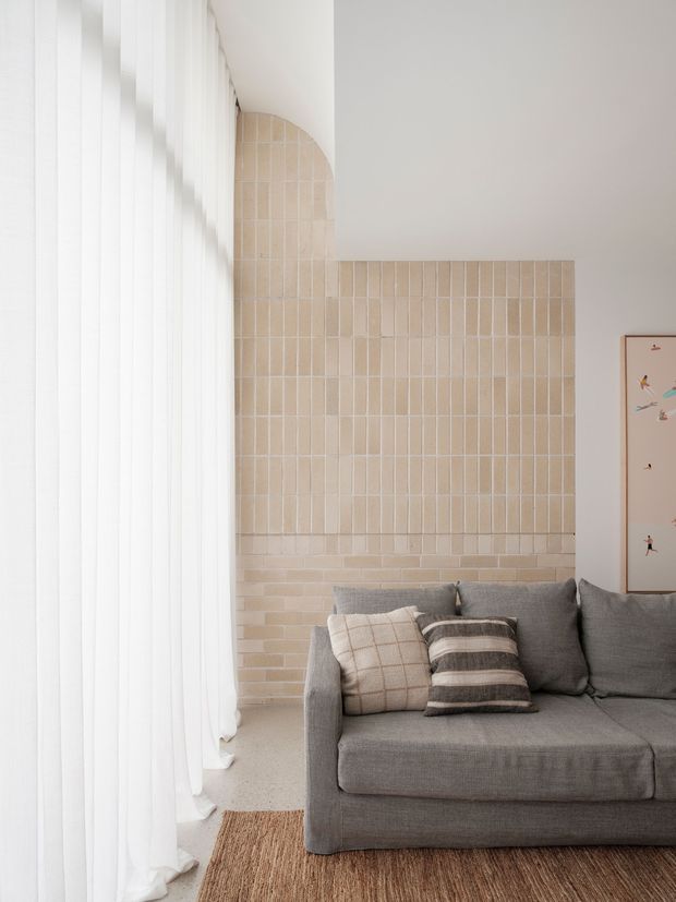 A bright, softly styled living room corner with a grey upholstered sofa, neutral cushions, full-height sheer white curtains, a warm woven rug, and a pale beige tiled feature wall with a curved upper detail. The space has a calm, contemporary and tactile aesthetic with natural neutral tones and abundant diffused daylight.