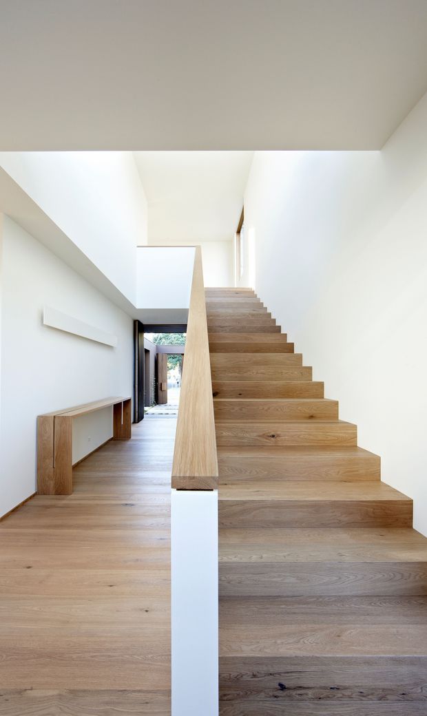 Bright minimalist interior hallway featuring a sculptural open timber staircase with broad oak-clad treads and a solid central balustrade. Pale timber flooring continues through the corridor, with white walls and ceiling, a simple built-in timber console, and a glazed doorway at the far end. The space is clean-lined and naturally lit.