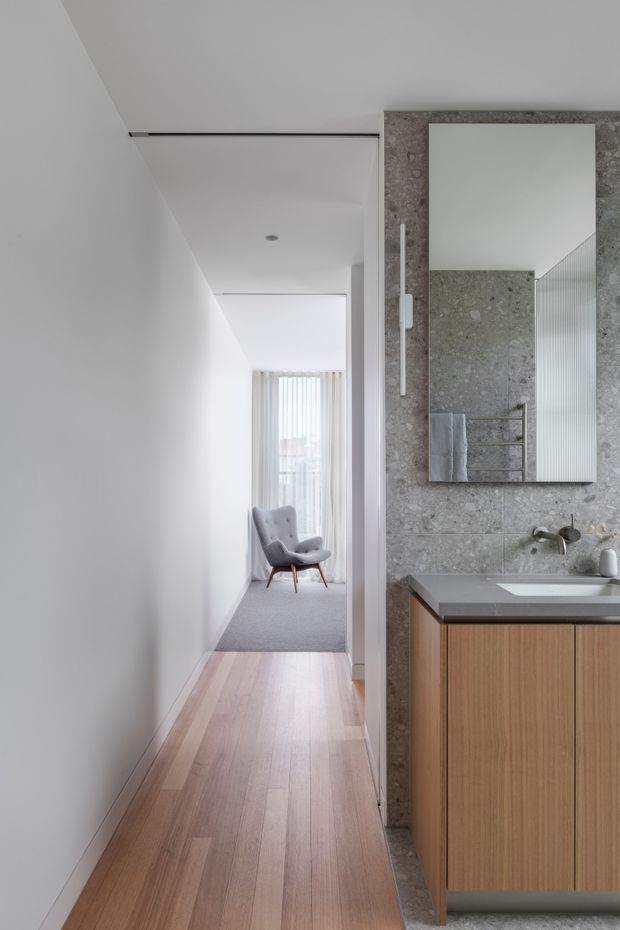 A narrow, minimalist residential hallway leading toward a softly lit sitting area, with a partially visible contemporary bathroom vanity on the right. The hallway has pale timber flooring, white walls and ceiling, recessed lighting, and a grey carpeted area near the armchair. The bathroom features a large grey stone-look tiled wall, frameless mirror, grey benchtop, and warm timber cabinetry.