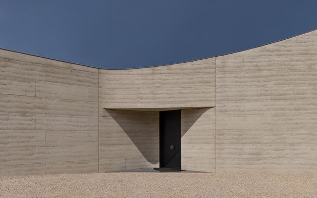 Minimalist exterior view of a curved, monolithic pale concrete or rammed-earth wall enclosing a small entrance. A deeply recessed dark door sits beneath a projecting angular canopy, creating strong geometric shadows. The scene is stark and sculptural, with a gravel foreground and clear blue sky.