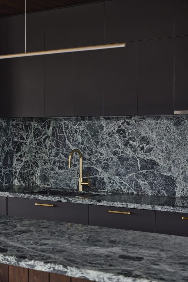 Close-up view of a dark, refined kitchen featuring near-black flat-panel cabinetry with slim brass-gold handles, a dramatic heavily veined dark stone splashback and benchtops, and a contrasting timber-lined ceiling. A minimalist linear pendant light and curved brass tap add understated luxury.