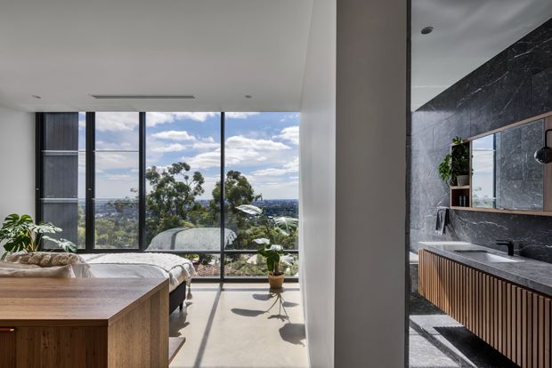 A contemporary open-plan interior showing a bedroom and bathroom separated by a partial wall. The bedroom has full-height black-framed glazing overlooking a leafy urban landscape, a timber bed base, and a large timber island or cabinet in the foreground. The adjacent bathroom features dark marble-look wall and floor surfaces, a floating timber vanity with vertical slatted doors, a dark benchtop, wall-mounted basin, black fixtures, and a large mirror. Natural light creates a bright, refined atmosphere.