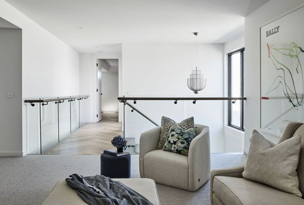 Bright contemporary upstairs landing or sitting area with a glass balustrade and black handrail overlooking a lower level. The space includes two upholstered armchairs, a small side table, a large ottoman or bench with a throw, pale carpet, light timber flooring in the hallway, a sculptural pendant light, and a large abstract artwork. A tall window provides natural light.