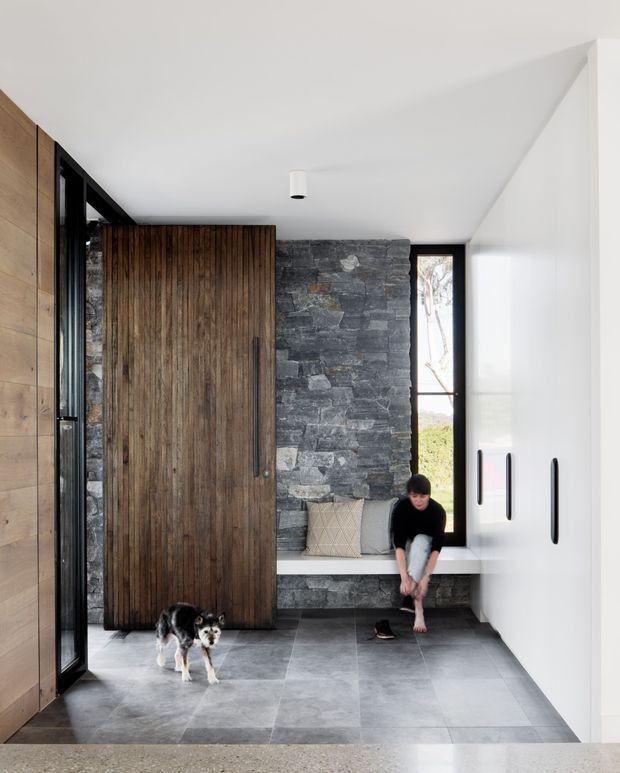 A bright, minimalist residential entryway with a large pivot-style timber door, dark stone feature wall, built-in white bench seat, tall black-framed glazing, and white built-in cabinetry. Large grey floor tiles and black hardware create a restrained contemporary material palette. A person and dog are present, with the entry opening toward an exterior view.
