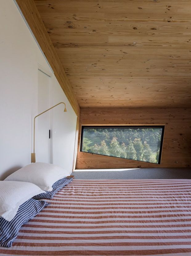 A compact, warmly detailed bedroom with a low angled timber-lined ceiling and matching timber wall around a large slanted picture window. A bed fills the foreground, dressed in a muted terracotta and white striped cover with layered white and blue-striped pillows. A slim brass wall-mounted reading light is fixed to the white wall beside the bed. The window frames a leafy wooded landscape, contributing natural light and an intimate cabin-like atmosphere.