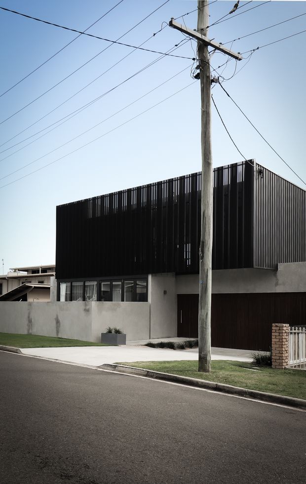 Street-facing view of a contemporary urban residence with a rectilinear form, smooth rendered walls, dark vertical slatted upper-level cladding, large windows, and an integrated dark garage door. The property sits behind a low rendered boundary wall with a small lawn and concrete driveway. A prominent timber utility pole and overhead wires occupy the foreground.