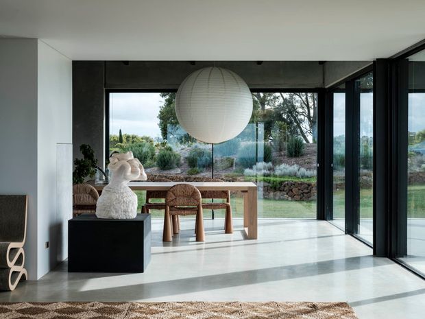 A bright, minimalist dining area with a simple timber dining table and woven chairs positioned beside expansive black-framed floor-to-ceiling glazing. A large spherical paper pendant hangs above the table, while a sculptural white object sits on a black plinth in the foreground. The polished pale floor, dark exposed ceiling beam, and landscaped garden visible beyond the glass create a calm contemporary interior.