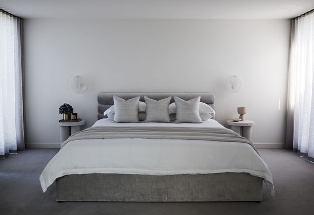 A serene, symmetrical contemporary bedroom with a large upholstered bed centred against a pale grey wall. Layered white and grey bedding, multiple cushions, soft wall-mounted lights, small bedside tables, and full-height sheer curtains create a calm, refined atmosphere. The room has carpeted flooring and abundant filtered daylight.