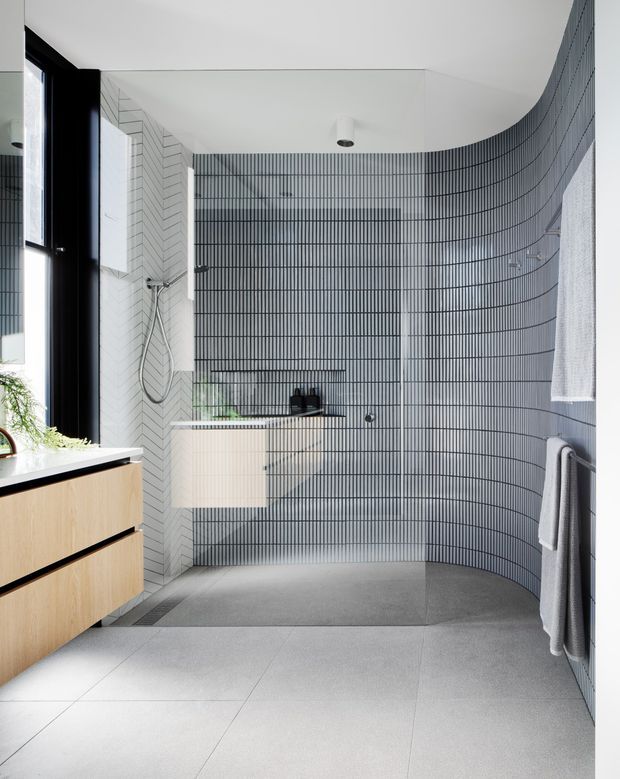 Contemporary bathroom with a curved walk-in shower enclosed by distinctive dark linear tiles. The space features pale grey large-format floor tiles, light timber floating vanity cabinetry, white geometric wall tiles, black-framed glazing, a wall-mounted basin, and minimal black fixtures. The bright, sculptural interior has a clean monochromatic palette with warm timber accents.