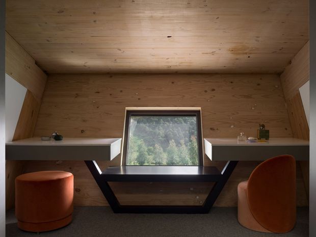 Compact timber-lined workspace or study nook with a fully clad wood ceiling, walls, and built-in side desks. A central window overlooks greenery, with a dark geometric trestle-style table or desk beneath it and two rounded burnt-orange upholstered stools/chairs. The warm, enclosed interior has simple contemporary detailing and subdued natural lighting.