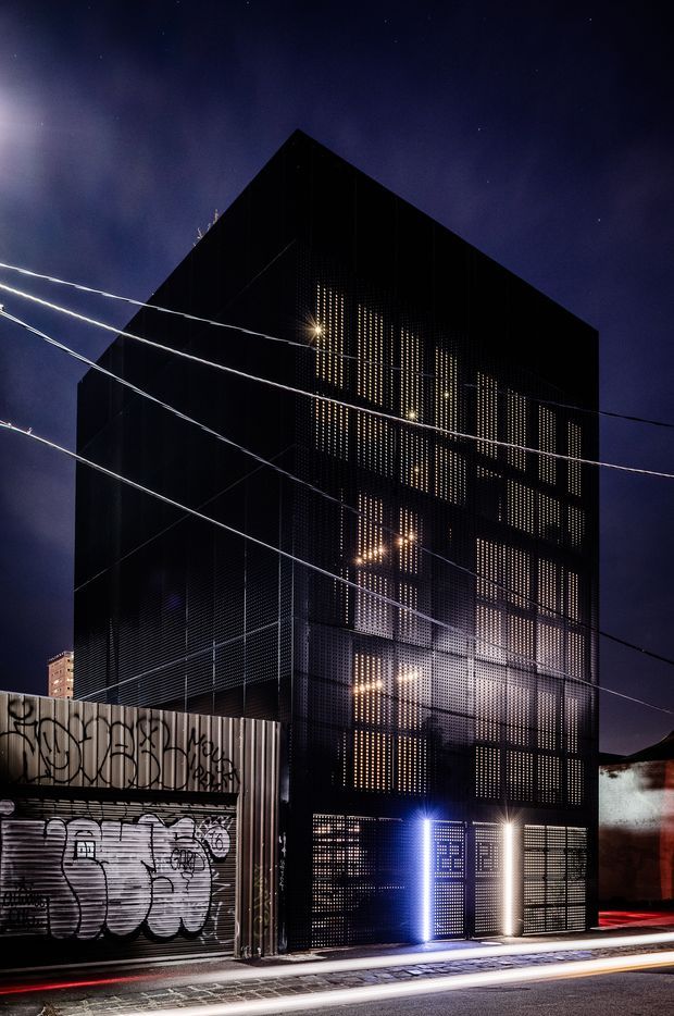 Night-time exterior photograph of a tall, narrow contemporary urban building with a sharply pitched or folded dark roof form and a perforated black metal facade. Warm interior light glows through the regular perforations, with brightly illuminated entry doors at street level. Graffiti-covered industrial roller doors and overhead utility wires frame the street-facing setting.