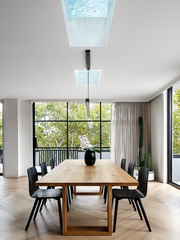 Bright contemporary dining room with a large natural timber dining table and black upholstered chairs, set against expansive black-framed glazing and a balcony railing with leafy trees beyond. Full-height sheer curtains, white walls, pale herringbone timber flooring, sculptural pendant lights, and indoor plants create an airy, refined interior.