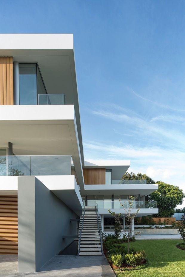 Contemporary multi-storey residential exterior with crisp white and grey geometric volumes, projecting balconies, clear glass balustrades, timber-look vertical screening, and a prominent external staircase. Landscaped lawn and planting surround the building under a clear blue sky.