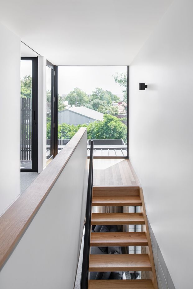 Bright contemporary stairway with open-riser timber stairs, slim black metal handrail, white walls, and large black-framed glazing at the upper landing. The windows overlook leafy suburban rooftops, while daylight creates an airy, minimal interior.
