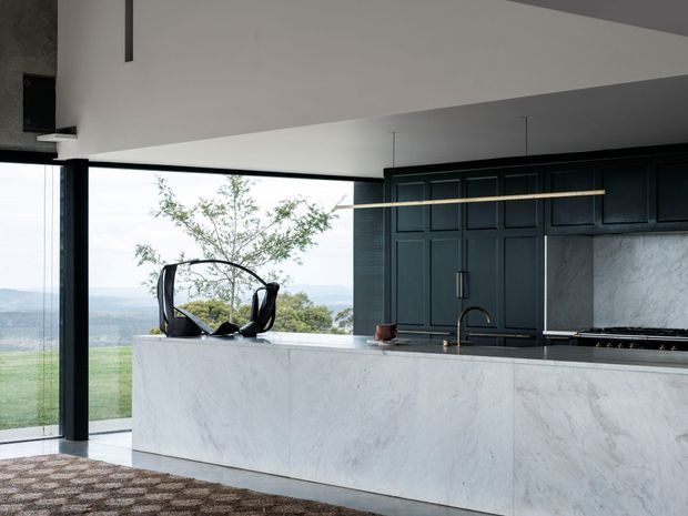A contemporary kitchen with a large white marble-look island, dark panelled cabinetry, integrated cooktop, brass-toned faucet and linear pendant light. Full-height glazing opens to a broad green and mountainous landscape, while a patterned rug adds warmth in the foreground. The room has a clean, refined, high-contrast composition with generous natural light.