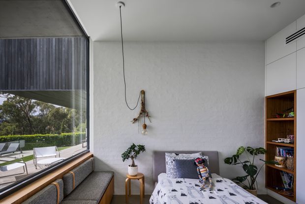 Compact contemporary bedroom with a built-in upholstered window seat beneath expansive floor-to-ceiling glazing. The room has a textured white wall, integrated timber shelving and cabinetry, a grey upholstered bed with patterned bedding, indoor plants, and a simple suspended pendant. A landscaped outdoor area is visible through the glazing.
