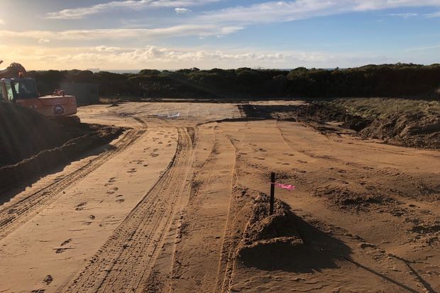 A broad sandy construction site with freshly graded ground, tire tracks, a small excavator at the left edge, and a marked survey stake in the foreground. Low coastal-looking vegetation lines the horizon under a partly cloudy sky.