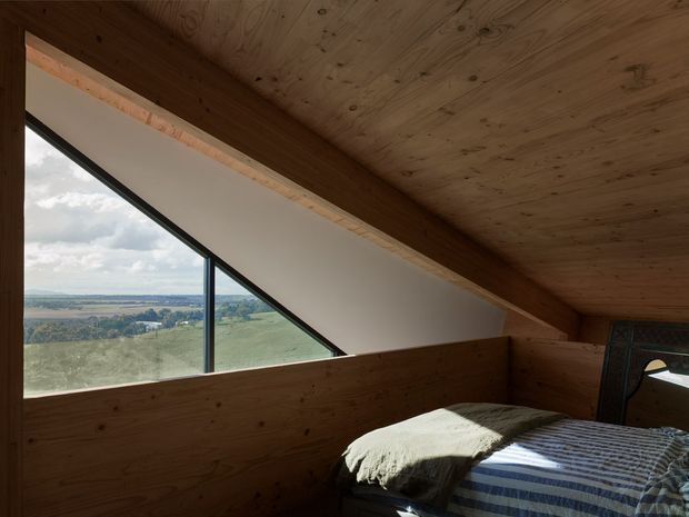 A compact bedroom tucked beneath a sloping timber-lined roof, with warm plywood or natural timber wall and ceiling surfaces. A large triangular window provides expansive views over open rural countryside. The room contains a bed with striped bedding and a dark framed mirror or opening at the side, creating a simple, rustic, cabin-like atmosphere.