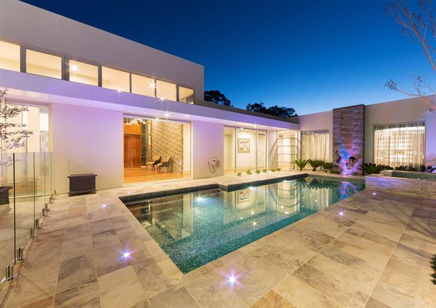 A contemporary two-storey residence arranged around a landscaped courtyard and illuminated swimming pool. The home features clean flat rooflines, extensive floor-to-ceiling glazing, pale rendered walls, a textured vertical stone-clad feature wall, and broad pale stone paving around the pool. Warm interior light and purple-blue landscape lighting create an upscale evening atmosphere.