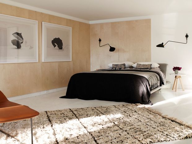 A warm, modern bedroom with pale timber wall panelling, a low upholstered bed dressed in black and grey bedding, and minimalist black wall-mounted reading lights. Framed monochrome artwork hangs on the timber wall, while a small white side table with flowers and a leather lounge chair add restrained furnishings. A large textured neutral rug covers much of the pale floor, and natural sunlight creates a bright, calm atmosphere.