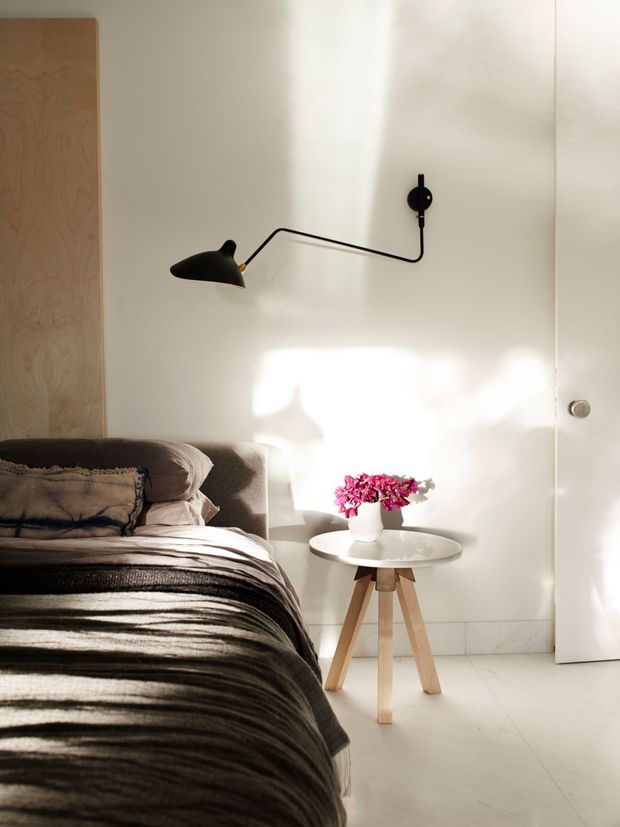 A softly lit bedroom corner with a partially visible bed dressed in layered neutral bedding, a light timber feature panel behind the bed, and a minimalist black articulated wall lamp. A small round white side table with timber tripod legs holds a vase of pink flowers. The room has pale walls, a light floor, and a simple white door, creating a warm, calm, contemporary atmosphere.