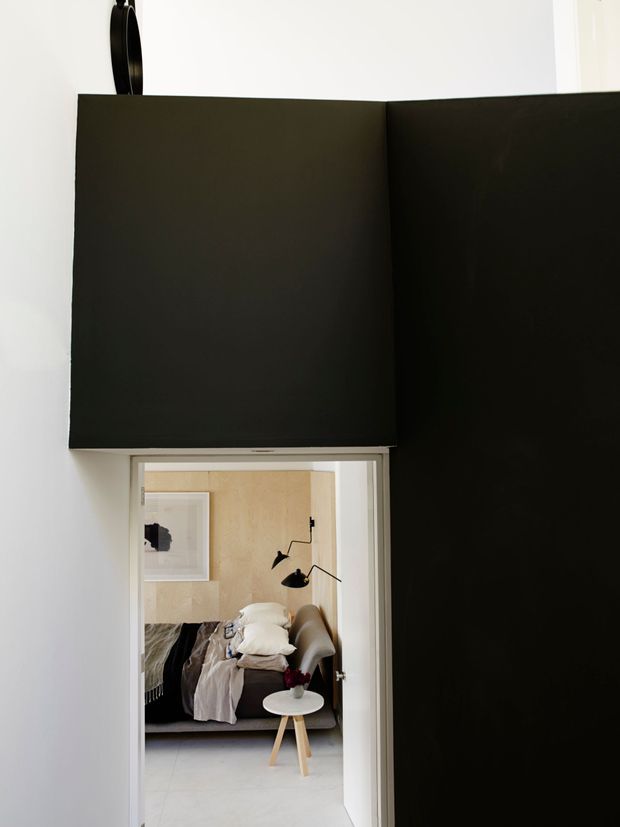 A minimalist interior view looking through a doorway into a compact bedroom. Large matte-black wall or door panels dominate the foreground, contrasting with white walls and a pale floor. The bedroom beyond features light timber wall panelling, a bed with layered neutral bedding, a grey upholstered armchair, a small round white side table, and a black articulated wall lamp.