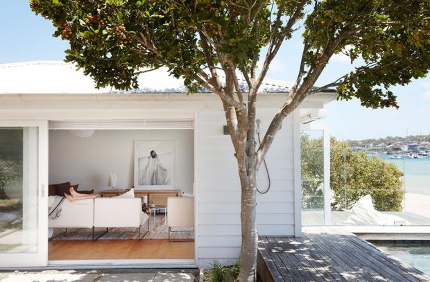 Bright coastal residence viewed from an exterior timber deck, with white horizontal weatherboard cladding, broad sliding glass doors, and a flat roofline. Through the open glazing is a light-toned living room with a sofa, armchairs, timber flooring, and artwork. A swimming pool occupies the foreground, while a sunlit waterfront harbour is visible beyond the property.