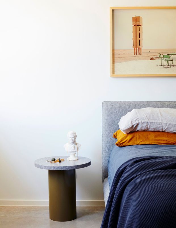 A bright, carefully styled bedroom corner with a grey upholstered bed, layered white, mustard-yellow and navy bedding, and a small round side table with a dark cylindrical base and pale stone-like top. A framed desert artwork hangs above the bed, while a sculptural bust and small objects sit on the bedside table. The room has pale walls, a light timber floor, and a clean contemporary aesthetic.