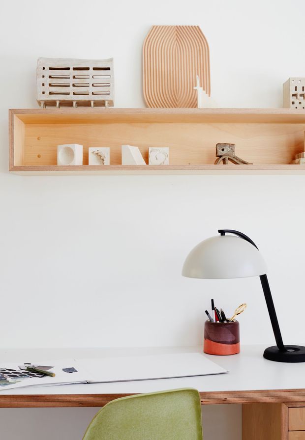 A bright, minimalist home workspace with a white desk, black-and-white task lamp, papers and drawing tools, and a green upholstered chair. A long open plywood wall shelf displays small sculptural objects and decorative pieces. The space has a clean, warm, modern studio aesthetic.