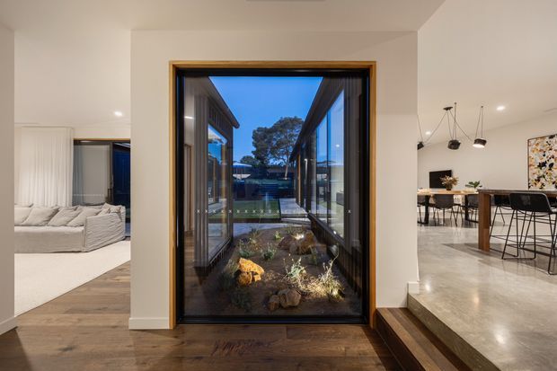 Interior view looking through a tall black-framed glazed opening onto a narrow landscaped courtyard or lightwell. The entry area has dark timber flooring and a small step up into a bright open-plan dining space with polished concrete flooring, a timber dining table, black chairs, pendant lighting, and artwork. A calm bedroom or living area with a light upholstered bed or seating is visible through an opening on the left.