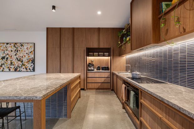 A warm, contemporary galley kitchen with extensive dark timber cabinetry, a large stone island, integrated appliance storage, and a long benchtop beneath a horizontal tiled or slatted backsplash. The space has polished concrete flooring, concealed under-cabinet lighting, open shelving with trailing plants, and a framed abstract artwork.